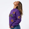 A woman wearing a purple crewneck sweatshirt with embroidered orange jack-o'-lanterns and a green witch hat, paired with denim shorts, photographed against a plain white studio background.