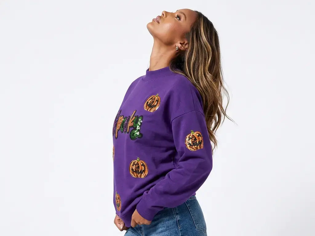 A woman wearing a purple crewneck sweatshirt with embroidered orange jack-o'-lanterns and a green witch hat, paired with denim shorts, photographed against a plain white studio background.