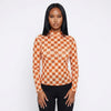 A woman modeling a long-sleeve mock neck top with an orange and beige checkerboard pattern featuring small pumpkin illustrations, paired with black pants, against a plain white studio background.
